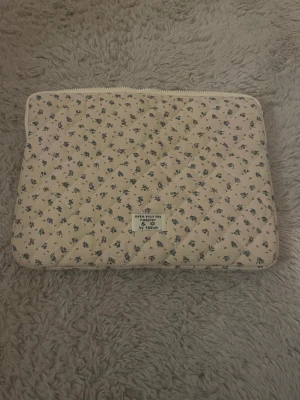 Blommig quiltad datorväska Kazum - Säljer en beige quiltad datorväska från Kazum med små blå blommor över hela väskan. Väskan har dragkedja upptill och är vadderad för extra skydd. Perfekt för dig som vill ha en söt och unik look till din laptop.