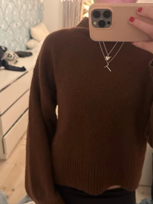 Brun stickad tröja med hög hals - Mysig brun stickad tröja med hög hals och ribbade muddar vid ärmslut och nederkant. Tröjan har en loose passform och är lite croppad, perfekt att styla med höga jeans. Materialet känns mjukt och värmande, idealisk för kyliga dagar.