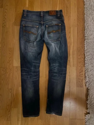 Nudie jeans - Säljer ett par riktigt feta jeans från Nudie med klassisk femficksdesign och schyssta slitningar. Jeansen har broderade bakfickor och en brun läderpatch med logga. Passformen är rak och materialet är robust denim.