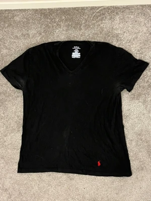 Svart v-ringad t-shirt Polo Ralph Lauren - Klassisk svart t-shirt från Polo Ralph Lauren med v-ringning och korta ärmar. Tillverkad i mjuk bomull och har den ikoniska röda logon broderad längst ner. Enkel och stilren modell som passar till allt. Tröjan är använd så slitage finns 