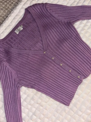 Lila ribbad kofta från AFJ - Säljer en lila croppad kofta från AFJ i storlek M. Koftan är ribbad och har långa ärmar samt v-ringning. Den stängs med pärlknappar framtill och är gjord i mjukt stickat material. 