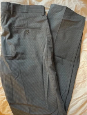 Grå kostymbyxor med smal rand - Snygga grå kostymbyxor med diskret smalrandigt mönster. Klassisk rak passform med knapp och ficka bak. Perfekta för en dressad look och passar till skjorta eller kavaj. Materialet känns mjukt och har en stilren finish.