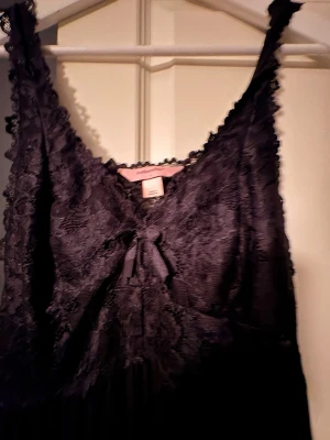 Svart spetsklänning från Hunkemöller - Supersnygg svart klänning från Hunkemöller med spets upptill och en söt rosett framtill. Klänningen har ärmlös design och v-ringad hals, med spetsdetaljer längs axelbanden och överdelen. Kjoldelen är slät och faller fint ner mot knäna.