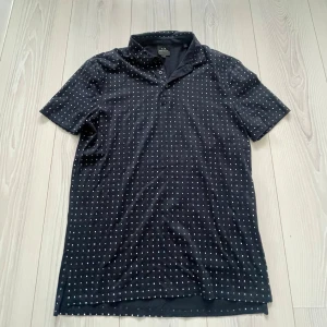 Svart prickig polotopp från Armani Exchange - Svart polotopp från Armani Exchange i storlek S med små vita prickar över hela plagget. Toppen har korta ärmar, klassisk krage och knäppning vid halsen. Skicka vid intresse! 