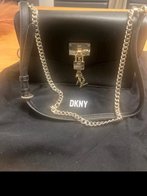 Svart handväska från DKNY med kedja - Snygg svart handväska från DKNY med silverfärgad kedja och detaljer. Väskan har ett lås framtill med DKNY-logga och hängande metallbokstäver. Tillverkad i skinn med stilren design och justerbar axelrem. Perfekt för dig som gillar klassisk och trendig look.