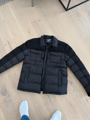 Svart pufferjacka från Brave Soul - Svart pufferjacka från Brave Soul, modell LDN. Jackan har en klassisk krage, två stora bröstfickor med lock och dragkedja framtill. Materialet är polyester och jackan har en stilren, mörk look som passar perfekt till kyliga dagar.