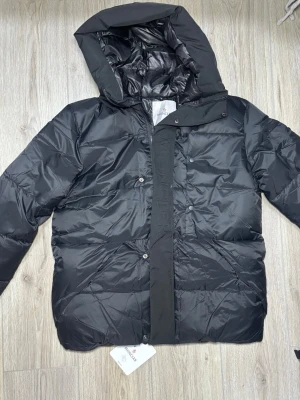 Svart Moncler pufferjacka med huva - Svart pufferjacka från Moncler med stor huva och tydlig logga på ärmen. Jackan har quiltad design, dragkedja och tryckknappar framtill. Materialet är glansig polyester och dunfyllning som håller dig varm under kalla dagar. Perfekt för dig som gillar stilren och exklusiv look.