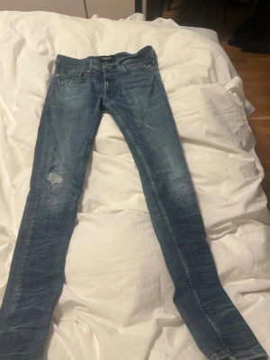Replay Jondrill jeans blå - Säljer ett par Replay Jondrill jeans i blå tvätt med rak ben passform. Jeansen har klassisk femficksdesign, ljusa slitningar och snygga detaljer på bakfickorna. Materialet är stretchigt jeans och midjan är normal. Perfekt för dig som gillar smalare jeans med cool look.