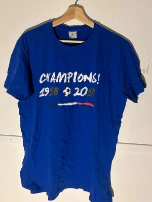 Blå Champions t-shirt från B&C Collection - Blå t-shirt från B&C Collection med trycket 'CHAMPIONS! 1998 2018' och en fotboll på framsidan. Snyggt motiv med färgstreck i grönt, vitt och rött. Baksidan har fransk text i vitt. Klassisk rund hals och korta ärmar. Perfekt för dig som älskar fotboll och vill sticka ut.