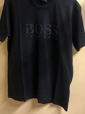 Svart t-shirt från Hugo Boss - Snygg svart t-shirt från Hugo Boss med stort BOSS-tryck på bröstet i ton-i-ton. Klassisk rund hals och korta ärmar. Tillverkad i mjuk bomull som känns skön mot huden. Perfekt för dig som gillar stilrena och trendiga plagg.