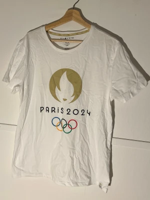 Vit Paris 2024 OS t-shirt - Säljer en officiell vit t-shirt från Paris 2024 OS med stort tryck framtill av OS-loggan och de klassiska olympiska ringarna i färg. T-shirten har rund hals och korta ärmar. Perfekt för dig som vill visa ditt stöd för spelen!