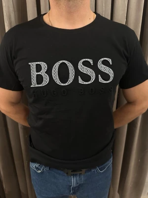 Svart BOSS t-shirt från Hugo Boss - Svart t-shirt från Hugo Boss med stort BOSS-tryck i vitt och svart på bröstet. Trycket har ett mönster med små BOSS-texter inuti bokstäverna. Rund hals och korta ärmar, tillverkad i mjuk bomull som känns skön mot huden. Perfekt för dig som gillar stilrena logotoplagg.