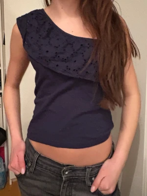 Mörkblå omlott topp med volang - Säljer en mörkblå omlott topp med volangdetalj och broderad struktur över axeln. Toppen är oneshoulder och croppad, vilket ger en snygg och modern look. Materialet känns som bomull och toppen har en tight passform som framhäver figuren.