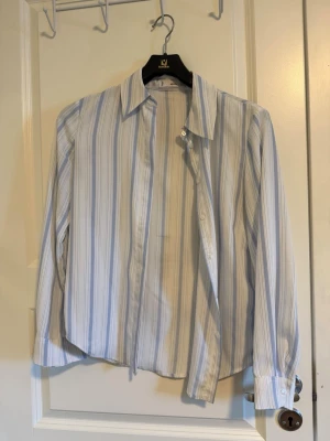Randig skjorta i blått och vitt - Snygg skjorta från mango med vertikala ränder i ljusblått och vitt. Klassisk krage, långa ärmar och knappar framtill. Perfekt för dig som gillar stilrena och fräscha plagg. Materialet är polyester.