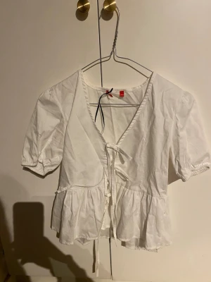 Vit omlottblus med knyt - Säljer en vit blus från RYVLS med puffiga korta ärmar och volang nedtill. Blusen har v-ringning och knyt framtill som ger en snygg siluett. Storlek 146/152. pris kan diskuteras