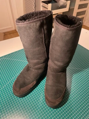 Svarta UGG boots med broderad detalj - Säljer ett par svarta UGG boots i mocka med grov sula och mjukt foder. Baksidan har en snygg broderad detalj i vitt och grått. Perfekta för kalla dagar och har en klassisk rund tå och platt sula. UGG-logga på hälen. Size 39