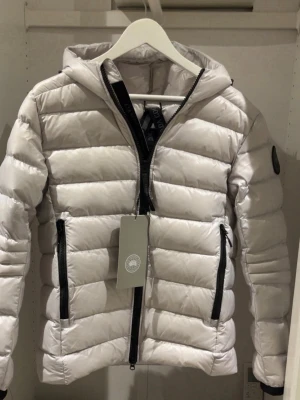 Ljusgrå pufferjacka från Canada Goose - Säljer en snygg ljusgrå pufferjacka från Canada Goose med huva och svarta dragkedjor. Jackan har ett stilrent quiltat mönster och logotyp på ärmen. Perfekt för kalla dagar och har en modern, loose passform.