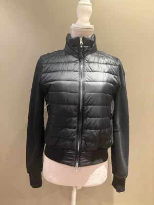 Svart pufferjacka från Parajumpers - Snygg svart pufferjacka från Parajumpers med quiltad framsida och hög krage. Jackan har långa ärmar i mjukt material, dragkedja framtill och en patch på ena ärmen. Perfekt för dig som vill ha en sportig och stilren look.