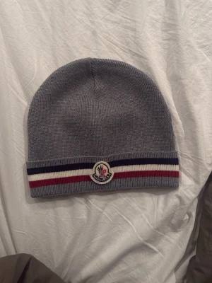 Grå Moncler mössa med ränder - Säljer en snygg grå mössa från Moncler med klassiska ränder i mörkblått, vitt och rött längs kanten. Mössan är stickad och har Moncler-loggan framtill. Perfekt för dig som vill ha en stilren accessoar med premiumkänsla. Pris kan diskuteras!!!