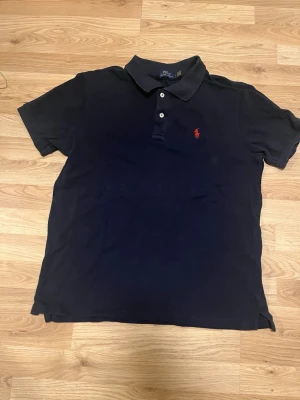 Svart pikétröja från Polo Ralph Lauren - Klassisk svart pikétröja från Polo Ralph Lauren i storlek XL. Tröjan har korta ärmar, krage och två vita knappar framtill. På bröstet finns det ikoniska röda Polo-logotypen broderad. Perfekt för en clean och stilren look.