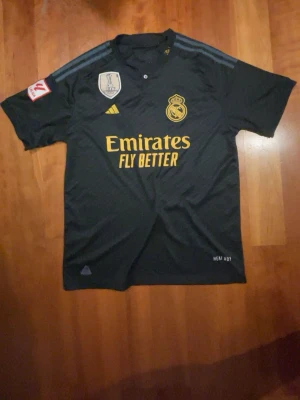 Real Madrid fotbollströja  - Bilderna visar en svart Real Madrid fotbollströja med gula detaljer och FIFA-märke, inte skor. Kan ej ge skobeskrivning.