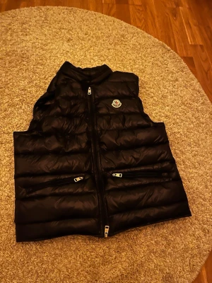 Svart dunväst från Moncler - Säljer en svart dunväst från Moncler med klassisk logga på bröstet. Västen har två dragkedjeförsedda fickor framtill och quiltad design som ger en snygg och sportig look. Perfekt att slänga över en hoodie eller tröja när det är kyligt ute.