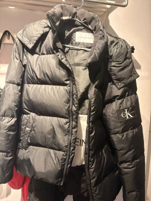 Svart pufferjacka från Calvin Klein Jeans - Säljer en svart pufferjacka från Calvin Klein Jeans med stor logga på ärmen och dragkedja framtill. Jackan har hög krage och breda kanaler för extra värme. Materialet är syntet och jackan har en clean, modern look.