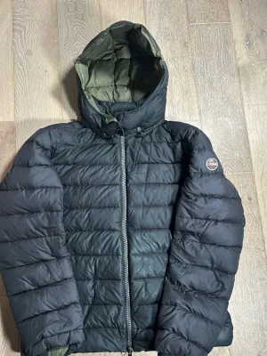 Svart pufferjacka från Colmar - Svart pufferjacka från Colmar med huva och grönt foder. Jackan har en rak passform, quiltad design och dragkedja framtill. Perfekt för kalla dagar och har en snygg badge på ärmen. Jackan är långärmad och har en clean look som funkar till många stilar.