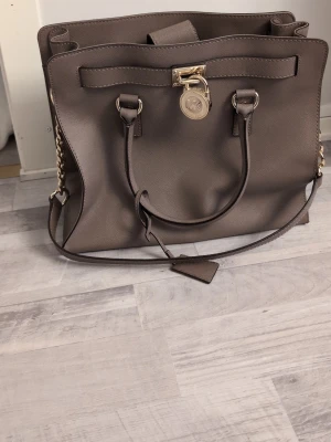 Brun handväska Michael Kors - Snygg brun handväska från Michael Kors med guldfärgade kedjedetaljer och MK-logga framtill. Väskan har både handtag och axelrem, samt ett dekorativt hänglås. Tillverkad i skinn med stilren, fyrkantig form och praktiska fack inuti. Perfekt för dig som gillar klassisk design med lyxig touch.