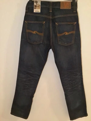 Mörkblå jeans från Nudie  - Säljer ett par mörkblå jeans från Nudie Jeans med klassisk orange söm och snygga detaljer på bakfickorna. Jeansen har en slim passform och perfekt för dig som gillar stilrena och moderna jeans. Jeansen är ändats testade och avända en gång då jag tog bort etiketten och inte kunde returnera. Pris ej hugget i stenen. Modellen är Slim Jim.