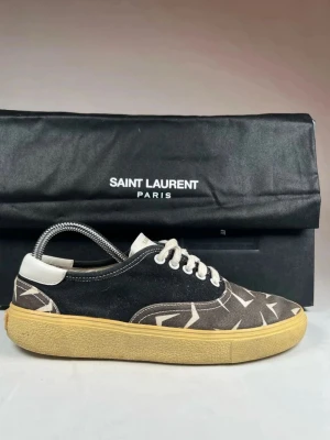 Saint Laurent sneakers - Snygga sneakers från Saint Laurent med svart och beige ovandel i bomull och grafiskt mönster. Skorna har vit snörning, rund tå och beige platt gummisula. Märkesdetalj på hälen och tungan. Perfekt för dig som gillar stilrena men unika sneakers.