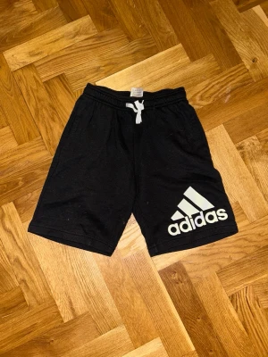 Svarta Adidas shorts med snörning - Svarta shorts från Adidas med vit logga på benet och vit snörning i midjan. Modellen är loose fit och har elastisk midja. Perfekta för träning eller chill. Materialet är mjuk bomull som känns skönt mot huden.