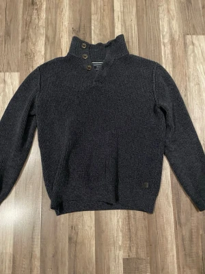 Mörkgrå stickad tröja Marc O'Polo - Säljer en mörkgrå stickad tröja från Marc O'Polo i storlek L. Tröjan har hög krage med tre snygga knappar och är tillverkad i twisted bomull. Den är långärmad och har en grovstickad struktur som ger en cool och avslappnad look.