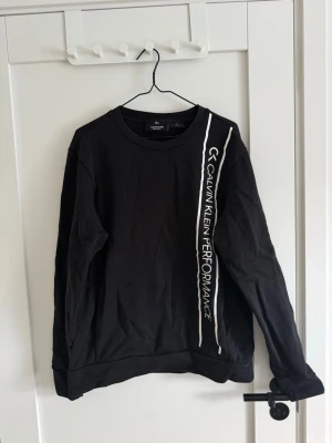 Calvin Klein tröja - Svart långärmad sweatshirt från Calvin Klein Performance med vit logotyp och tryck längs ena sidan framtill. Tröjan har rund hals och är gjord i mjuk bomull, perfekt för en sportig och avslappnad stil.
