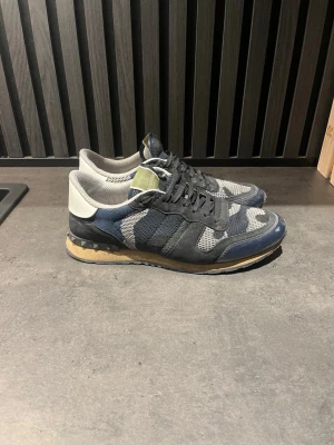 Valentino Rockrunner sneakers i blått - Säljer ett par Valentino Rockrunner sneakers med blå, grå och vita camouflage-detaljer. Skorna har snörning, rund tå och platt sula med nitar på hälen. Materialmix av mocka, mesh och skinn. Snygg kontrast mellan färgerna och coola detaljer på sulan.