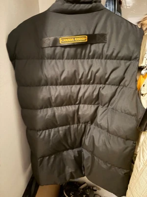 Svart dunväst från Canada Goose - Säljer en svart dunväst från Canada Goose med klassisk logotyp på bröstet och broderad etikett på ryggen. Västen har hög krage, dragkedja och tryckknappar framtill samt praktiska fickor. Perfekt för kyliga dagar och har en stilren, quiltad design.