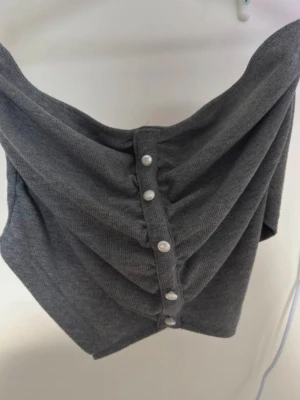Mörkgrå bandeau tubtopp med pärlknappar - Snygg mörkgrå bandeau tubtopp med ribbad struktur och dekorativa pärlknappar framtill. Toppen är axelbandslös och har en draperad effekt som ger en cool och trendig look. Perfekt att styla med jeans eller kjol för en modern vibe.