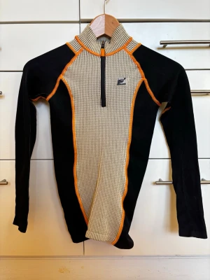 Randig oanvänd långärmad tröja från Iets Frans Urban Outfitters - Säljer en långärmad tröja från Iets Frans Urban Outfitters med randigt mönster i beige och svart, orange sömmar och half zip framtill. Tröjan har hög krage och är tillverkad i ett stickat material som känns både sportigt och retro. Oanvänd!