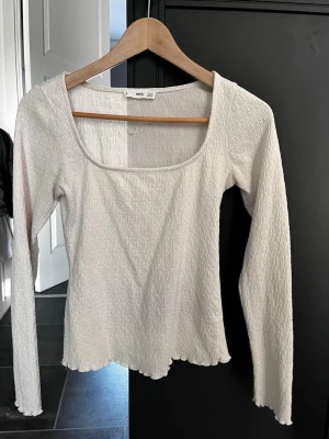 Vit långärmad topp från Mango - Säljer en vit långärmad topp från Mango med fyrkantig urringning och lätt ribbad struktur. Toppen har vågig kant nedtill och är tillverkad i ett mjukt syntetmaterial. Perfekt att styla med jeans eller kjol för en clean look.