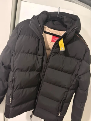 Svart pufferjacka Parajumpers S - Svart pufferjacka från Parajumpers i storlek S. Jackan har beige insida, huva, två dragkedjefickor och en gul detalj vid halsen. Tillverkad i polyester och dun, perfekt för kalla dagar. Snygg och funktionell design med ribbade muddar och säkerhetsficka.
