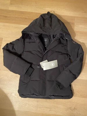 Svart pufferjacka från Canada Goose - Svart pufferjacka från Canada Goose med huva, ribbade muddar och diskret logotyp på ärmen. Jackan har dragkedja och knappar framtill, samt två fickor. Perfekt för kalla dagar och har en clean, modern look.