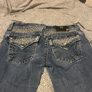 Miss Me jeans med paljettdetaljer - Snygga blå jeans från Miss Me med glittriga paljettdetaljer och pärlknappar på fickorna. Märkets emblem i skinn på bakfickan. Jeansen har låg midja och bootcut, med slitningar och coola detaljer som ger en unik look. Lite slitna (sista bilderna) originalpris 2500kr. 1250kr +frakt