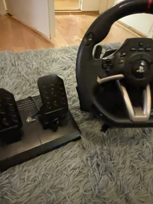 Hori Racing Wheel Apex - Hori Racing Wheel Apex med pedaler, kompatibel med PS4 och PS3. Perfekt för racingspel, med PlayStation-knappar och justerbara inställningar. Produkten ser ut att vara i gott skick utan synliga defekter eller större slitage. Komplett med kablar och fästen.