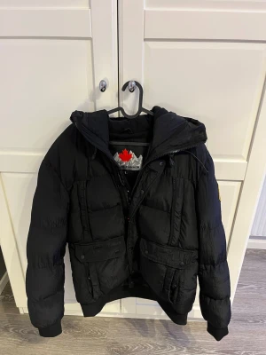 Svart pufferjacka från Zavetti Canada - Svart pufferjacka från Zavetti Canada med huva och stora fickor framtill. Jackan har quiltad design, ribbade muddar och dragkedja. Insidan har tryckt logga och mönster. Perfekt för kalla dagar och riktigt snygg streetstil. Storleken är L men sitter som M/S. Jackan är endast använd ett fåtal gånger.