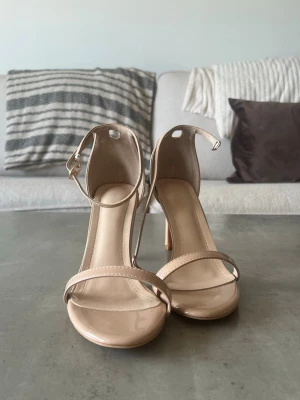 Beige klackar / sandaler med smal rem - Snygga beige sandaler med hög klack och smala remmar över tårna samt runt ankeln. Skorna har en rund tå och är tillverkade i glansigt syntetmaterial. Perfekta för dig som gillar stilrena och minimalistiska skor med en elegant touch. Storlek 40 men passar 39/38 