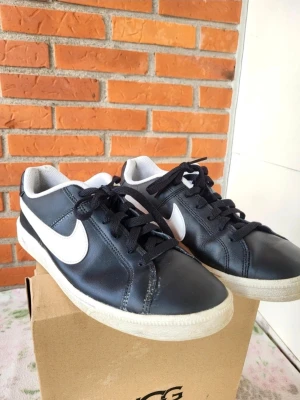 Svarta Nike sneakers med vit swoosh 42 - Säljer ett par svarta Nike sneakers med vit swoosh på sidorna och vit sula. Skorna har snörning, rund tå och lågt skaft. Ovandelen är i skinn och insidan är fodrad med vit mesh. Perfekt för dig som gillar klassisk streetstil.