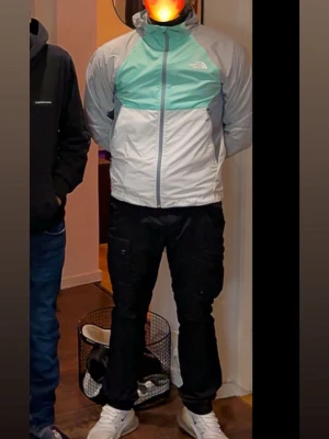 Mintgrön och grå vindjacka från The North Face - Fräsch vindjacka från The North Face i mintgrönt, grått och vitt. Jackan har huva, hel dragkedja och två sidofickor med dragkedja. Lätt och smidig design i syntetmaterial, perfekt för blåsiga dagar. Logga på bröstet och cool färgblockad look.       Väldigt sällsynt windbreaker från märket north face som använts väldigt snålt då den är limited edition , tagit väl hand om den som ni kan se så den är i princip så gott som ny passar s/m 