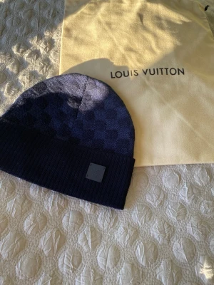 Mörkblå LV mössa - Snygg mörkblå mössa från Louis Vuitton med diskret rutigt mönster och ribbad kant. Märke i läder framtill och tillverkad i 100% ull. Perfekt accessoar för dig som gillar exklusiva detaljer och stilren design.