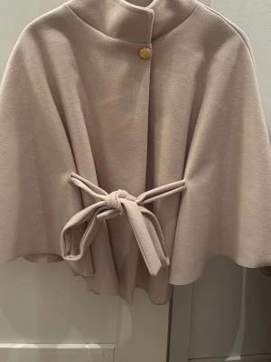 Beige poncho med knytband - Säljer en beige poncho med snyggt knytband i midjan och en guldig knapp vid halsen. Jackan har en mjuk och lyxig känsla, med en elegant och flowy siluett. Perfekt för dig som gillar stilrena och klassiska plagg med en modern twist.
