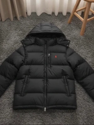 Svart dunjacka från Polo Ralph Lauren - Svart dunjacka från Polo Ralph Lauren med röd broderad logga på bröstet. Jackan har huva, två sidofickor och RL-detalj på dragkedjan. Puffad design och långärmad, perfekt för kalla dagar. Materialet är polyester och dun, med en clean och stilren look. Pris går absolut o diskutera bara o höra av sig 